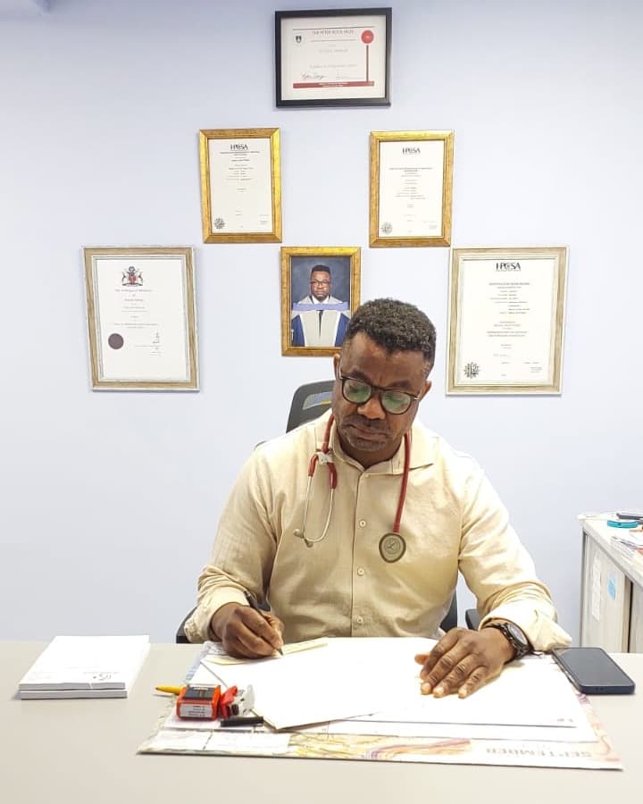 Dr Makanda writing patient notes at his consulting-room desk, wearing a stethoscope, with his framed medical qualifications and HPCSA registration certificates on the wall behind